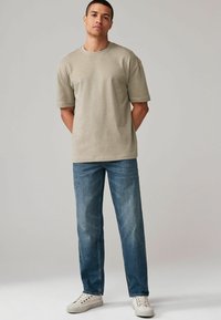 Tricou din bumbac de culoare bej deschis, cu guler rotund și mâneci scurte, asortat cu blugi albastri cu croială dreaptă și adidași albi.