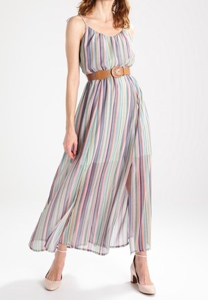 Kobieta ubrana w bezrękawnikową, wielokolorową, pionowo paskowaną sukienkę maxi z brązowym paskiem i beżowymi szpilkami, stojąca na tle białego tła.