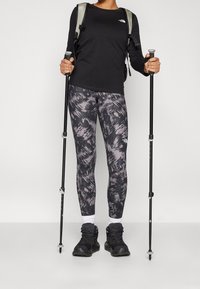 Sort langærmet skjorte, mønstrede leggings i nuancer af grå, robuste vandrestøvler og trekkingstænger. Rygsækken er sølvfarvet med sorte accenter.