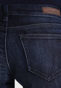 Los jeans de denim oscuro presentan un diseño ajustado con un suave desgastado. El bolsillo trasero tiene una solapa puntiaguda y una etiqueta de cuero marrón está cosida en la cinturilla.