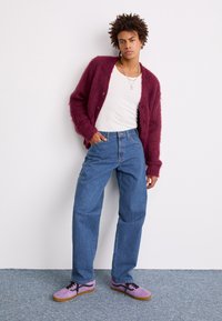 Burgunderfarget, fuzzy cardigan, hvit tettsittende tank top, blå høyhuskede jeans og lilla sko med svarte lisser. Står mot en hvit bakgrunn.
