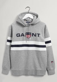 Felpa grigia con strisce bianche e blu navy, testo "GANT" e logo a scudetto, appesa a una gruccia bianca contro uno sfondo di muro chiaro.