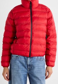 Rød vinterjakke med høj krave, med vandret quiltede sektioner, en sort frontlynlås og sidelommer. Båret over blå denimjeans.