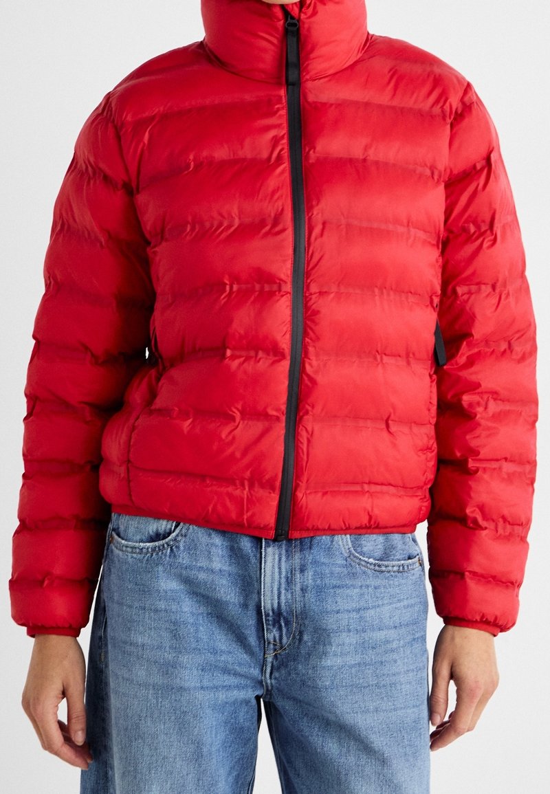 Rød vinterjakke med høj krave, med vandret quiltede sektioner, en sort frontlynlås og sidelommer. Båret over blå denimjeans.