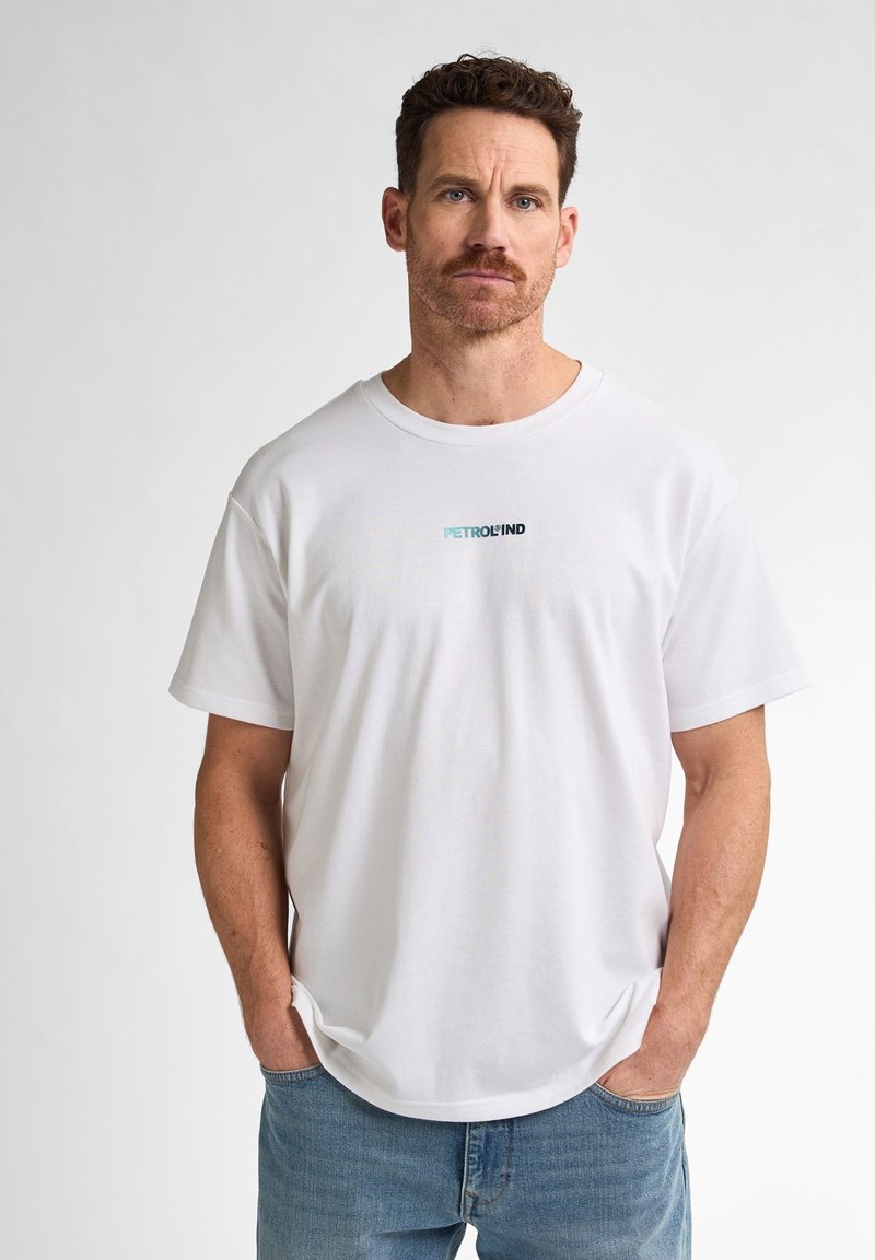 Un tricou alb din bumbac, cu mâneci scurte, decolteu rotund și un mic logo "PETROLIND" în teal și negru pe piept. Sunt vizibile blugi din denim.