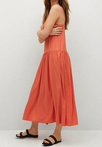 Robe sans manches corail avec une taille froncée et une jupe fluides. Le tissu a une finition texturée. Associée à des sandales noires.