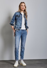 Jeansjacka med ljusare blåtvät, knäppning med knappar och framfickor. Vit t-shirt under. Bleka blå jeans och vita sneakers.