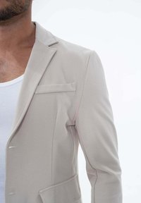 Blazer beige clair en tissu lisse, avec une poche poitrine simple et un col cranté, présentant une silhouette ajustée.