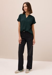 Mørkegrøn kortærmet bluse i struktureret stof, V-udskæring; parret med højtaljede sorte jeans og beige sneakers med grønne detaljer.