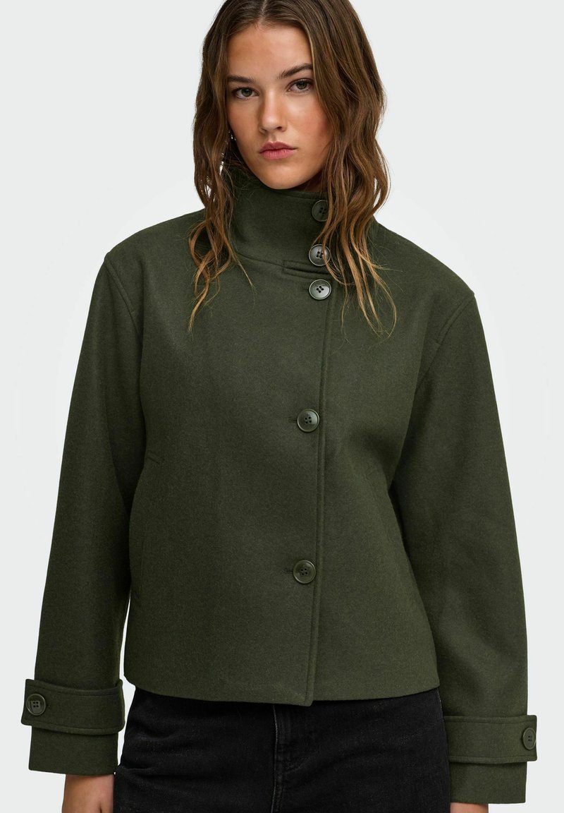 Jeune femme portant une veste boutonnée vert foncé à col montant, manches longues et pantalon noir, face à un fond blanc.