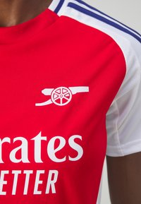 Červený fotbalový dres s bílými akcenty, se znakem děla a textem "Emirates", vyrobený z hladké, prodyšné látky.