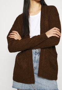 Cardigan marrone lavorato a maglia con design aperto, con polsini a coste, due tasche frontali, indossato sopra una t-shirt bianca e una gonna di jeans. Texture morbida.