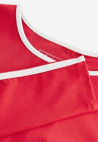 Czerwony top z materiału z okrągłym dekoltem i kontrastującymi białymi wykończeniami. Gładka tekstura z przeszytymi brzegami i widoczną małą metką.