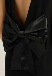 Vestido negro con un profundo escote en V en la espalda y un gran detalle de lazo de satén en la cintura, que resalta la suave textura del tejido y el diseño elegante.