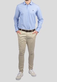 Camicia azzurra a maniche lunghe con bottoni, chinos beige, cintura marrone e sneakers bianche, in piedi contro uno sfondo grigio.