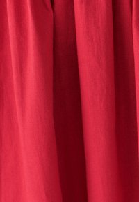 Roter Stoff mit weicher Textur und vertikalen Falten. Das Gewebe wirkt leicht strukturiert, was auf ein leichtes Material hinweist.