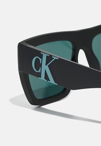 Ochelari de soare negri cu lentile de nuanță teal, având un logo "cK" de un albastru deschis bine definit pe lateral. Design unghiular, finisaj mat și rame late.
