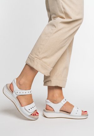Feet wearing white sandals with metal studs and beige cuffed pants on a white background, one foot slightly raised.