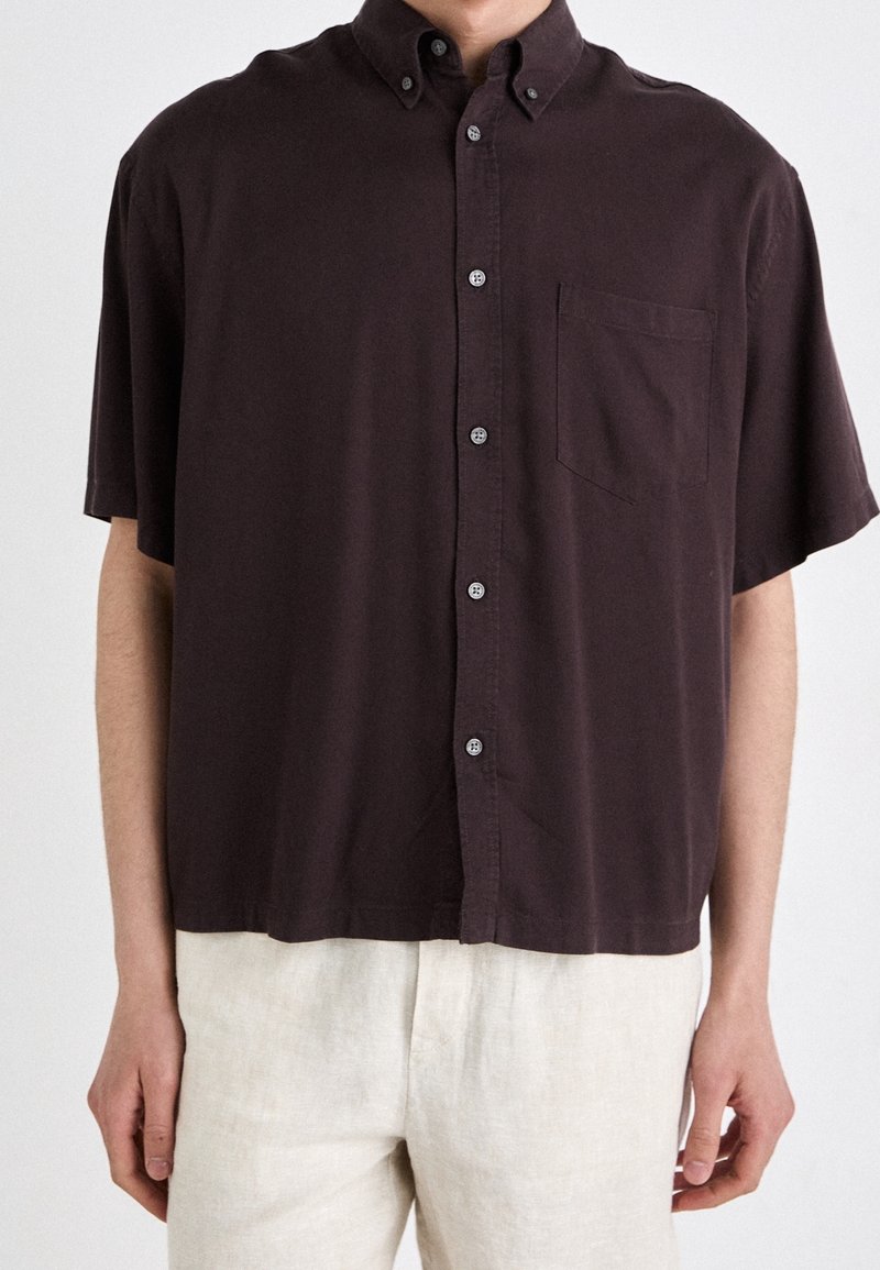 Camisa de manga corta con botones en algodón marrón oscuro. Cuenta con un bolsillo en el pecho y un corte relajado con detalles sutiles en el cuello.