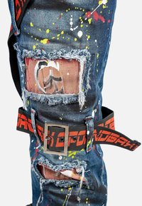 Pantalons en denim usés avec une couleur bleue et des motifs de éclaboussures de peinture en rouge, jaune et blanc. Comprend des patchs aux genoux effilochés et une ceinture noire.