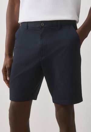 Mand iført mørkeblå buttoned shorts med hænderne i lommerne og en hvid skjorte synlig i taljen.