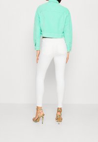 Miętowozielona krótka dżinsowa kurtka, białe dopasowane spodnie oraz wielokolorowe sandały na obcasie z paskami. Widok z tyłu ukazujący szczegóły stroju.