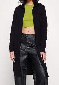 Czarny długi kardigan z kapturem, prążkowanymi mankietami i bocznymi kieszeniami, zestawiony z zielonym krótkim topem i czarnymi skórzanymi spodniami, tworząc warstwową stylizację.