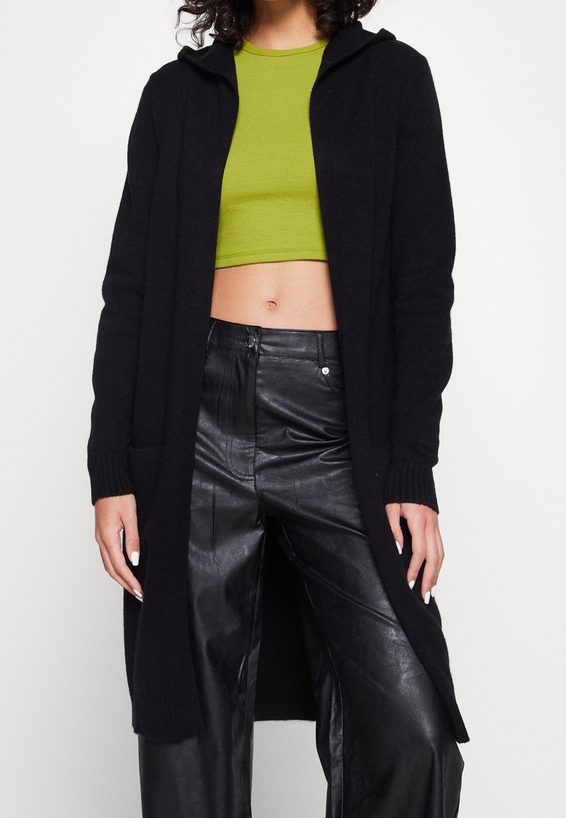 Czarny długi kardigan z kapturem, prążkowanymi mankietami i bocznymi kieszeniami, zestawiony z zielonym krótkim topem i czarnymi skórzanymi spodniami, tworząc warstwową stylizację.