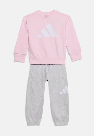 Survêtement enfant avec un sweat-shirt rose à manches longues et un pantalon de jogging gris clair, tous deux arborant un grand logo blanc de la marque.