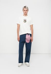 White t-shirt with colorful graphic design, loose blue denim trousers, and a pink cap hanging from the waistband. Footwear: white sneakers.