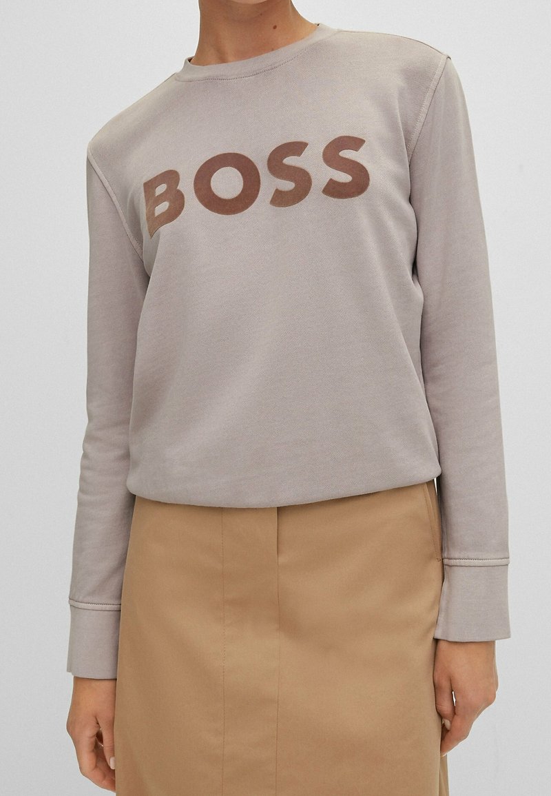 Ljusgrå sweatshirt med rund halsringning, prydd med ordet "BOSS" i brunt över bröstet. Mjuk textur, långa ärmar, enkel design.