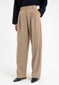 Beige wijde broek met hoge taille, plooien en riemlussen. Gemaakt van gladde stof, gecombineerd met zwarte schoenen met een puntige neus.