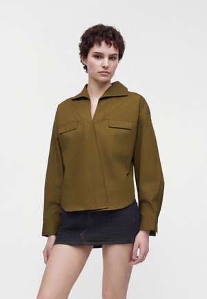 Jeune personne aux cheveux courts et bouclés portant une chemise à manches longues vert olive et une mini-jupe en jean foncé sur un fond uni.
