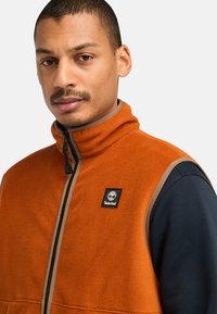 Fleece vest in bright orange with a high collar, zip closure, and a black logo patch. Lightweight, textured material with beige accents.