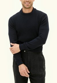 Marineblauer Pullover mit strukturiertem Strickmuster, geripptem Ausschnitt und Bündchen. Kombiniert mit schwarzen Hosen. Arme vor der Brust verschränkt.