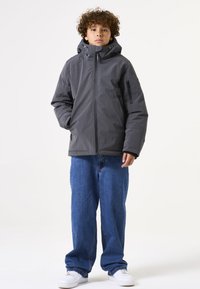 Chaqueta gris aislante con un alto cuello, cremallera frontal y bolsillos laterales, combinada con jeans azules holgados y zapatillas blancas.