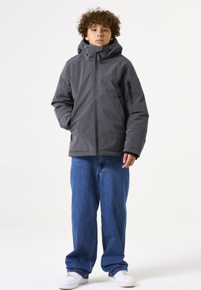 Chaqueta gris aislante con un alto cuello, cremallera frontal y bolsillos laterales, combinada con jeans azules holgados y zapatillas blancas.