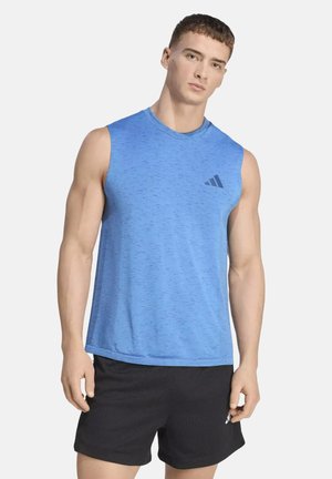 Jeune homme portant un débardeur de sport bleu et un short noir, debout devant un fond blanc uni.