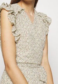 Kukkakuviollinen mekko, jossa on kermaan vivahtava kangas, röyhelöiset hihat, v-pääntie ja solmittava vyötärönauha, joka korostaa mekon naisellista muotoilua.