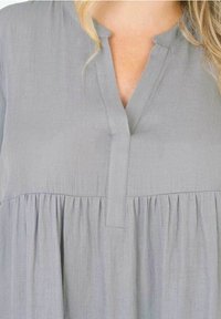 Světlá šedá blůza s výstřihem do V, nařasením v pase a měkkou, strukturovanou tkaninou. Obsahuje zapínání na knoflíky na přední části.