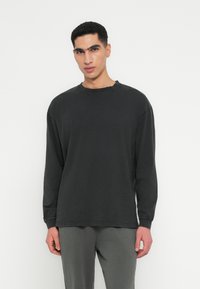 T-shirt nero a maniche lunghe in tessuto morbido, colletto rotondo, vestibilità rilassata, con dettagli di cucitura sottili. Abbinato a pantaloni grigi.