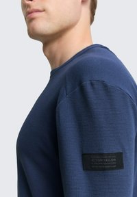 Marineblaues, strukturiertes langärmliges Hemd mit Rundhalsausschnitt und einem Etikett an der linken Ärmeln, das Markeninformationen enthält.