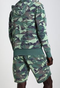 Conjunto de sudadera con capucha y pantalones cortos de patrón de camuflaje en verde y azul marino. Hecho de material suave con capucha. Presenta un ajuste casual y un dobladillo deshilachado en los pantalones cortos.