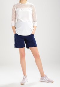 Biała bluzka z koronkowymi detalami na rękawach, w połączeniu z granatowymi szortami i jasnoróżowymi butami sportowymi z białymi paskami.