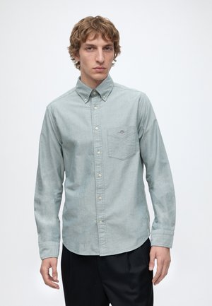 Jeune homme aux cheveux courts et bouclés, portant une chemise gris clair à boutons et un pantalon noir, debout devant un fond blanc.