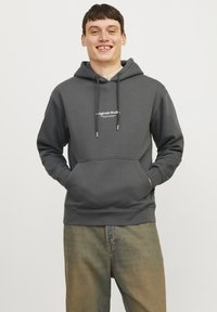 Grå hettegenser med kengurulomme, snøring på hetta, og en hvit logo på brystet, kombinert med lyse avslappede jeans.