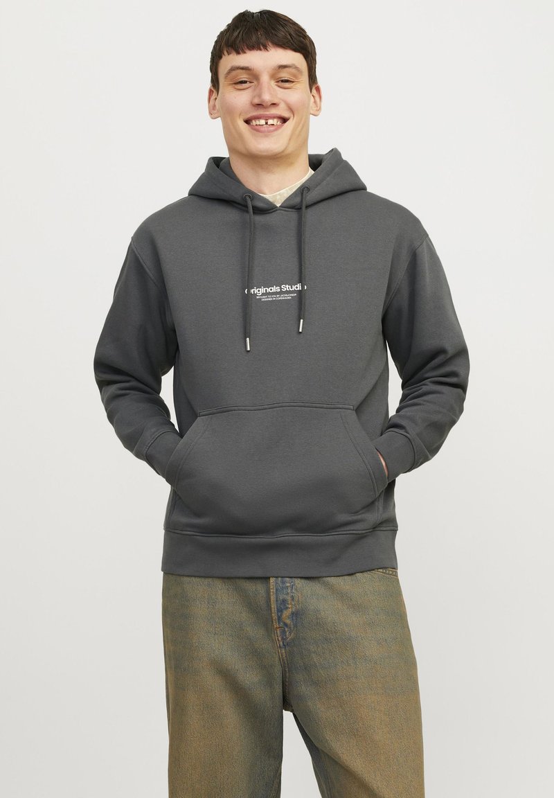 Grå hettegenser med kengurulomme, snøring på hetta, og en hvit logo på brystet, kombinert med lyse avslappede jeans.