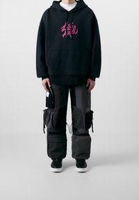 Felpa nera con cappuccio e grafica rosa, abbinata a pantaloni cargo con molteplici tasche e dettagli neri. Scarpe da ginnastica nere completano il look.