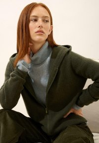 Sweat à capuche en tricot côtelé vert foncé, avec zip sur le devant, superposé à un col roulé bleu clair. Modèle assis, portant un pantalon en velours côtelé.