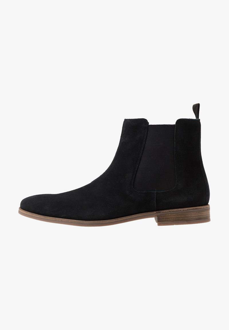 Stivale Chelsea in suede nero con pannelli elastici laterali e un tassello. Presenta una punta arrotondata e una suola in contrasto di colore marrone chiaro.